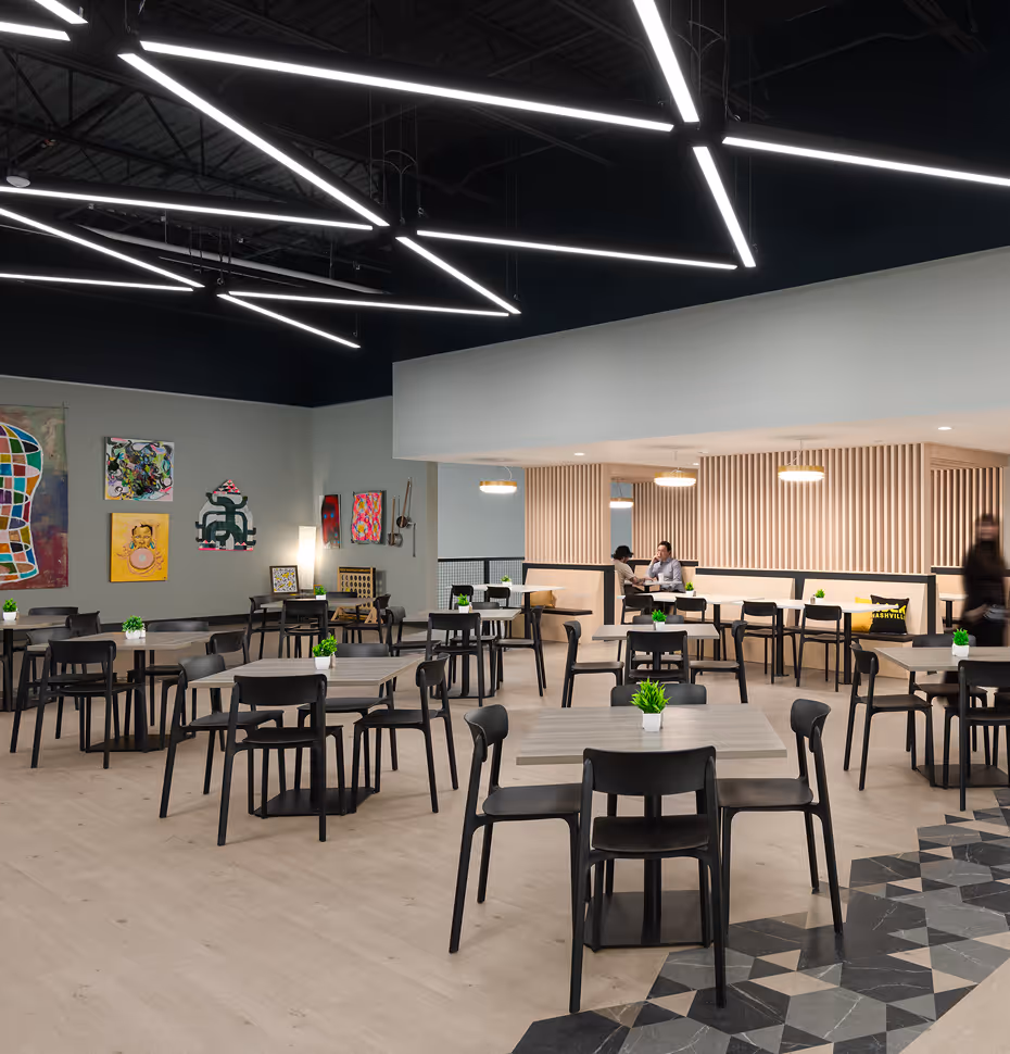 Modern cafeteria with multiple gray tables and black chairs, small green plants on each table, artistic wall decor, and unique geometric ceiling lights.