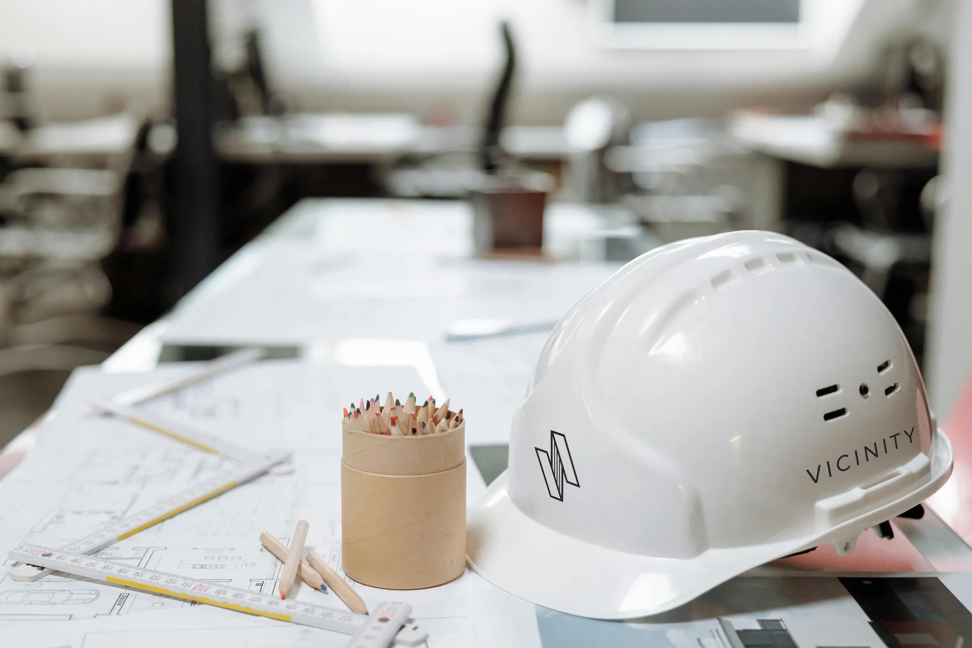 White hard hat labeled 'VICINITY' placed on architectural blueprints with a container of colored pencils and measuring tools nearby.