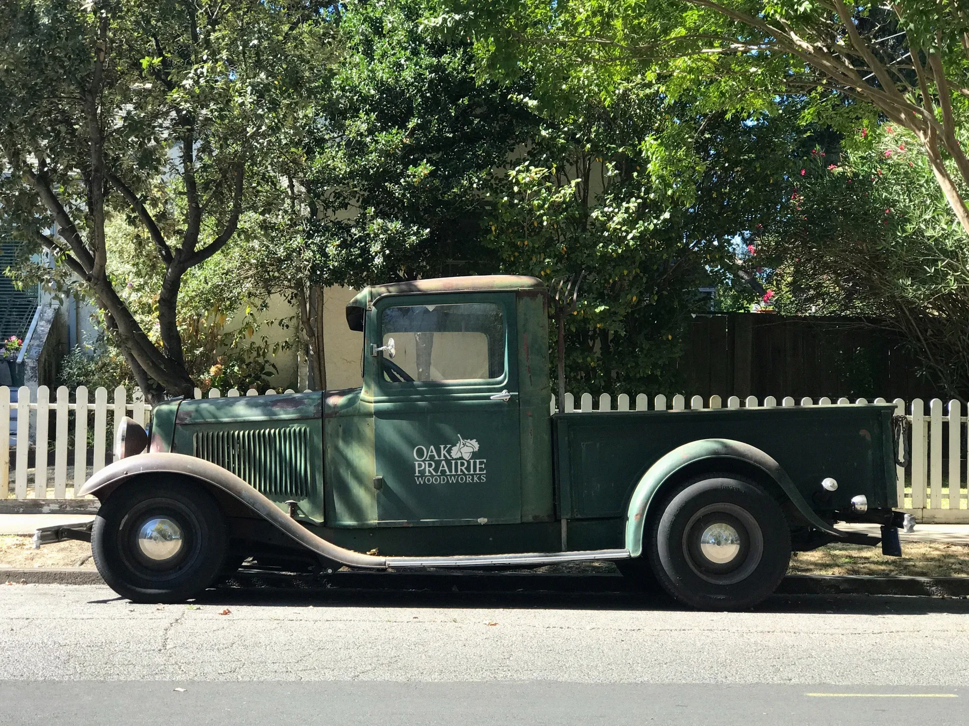 Vintage green pickup truck with Oak Prairie Woodworks logo parked in front of a white picket fence and trees.