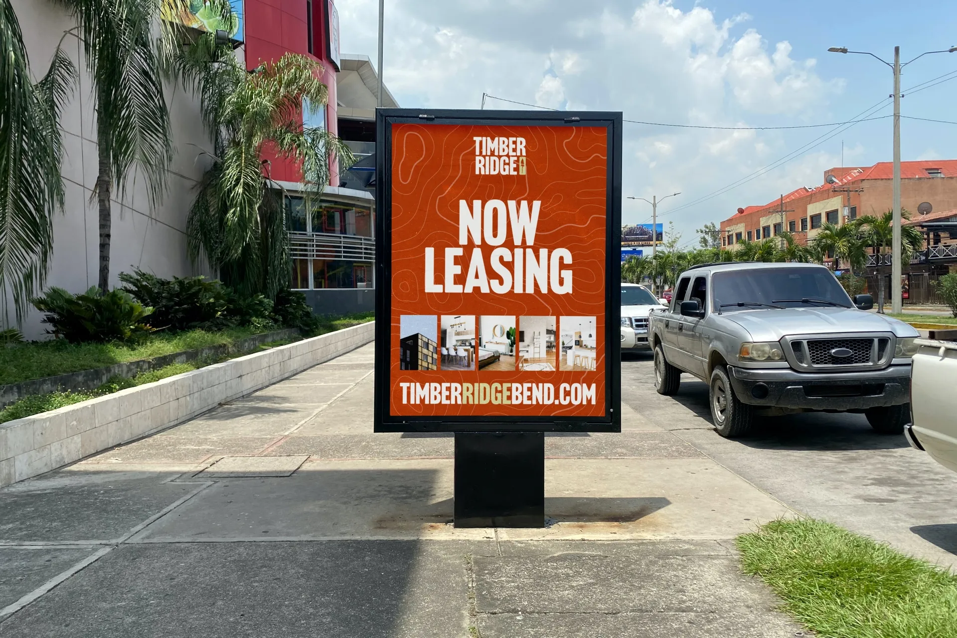 Outdoor sidewalk sign with orange background displaying "Timber Ridge Now Leasing" and images of modern apartment interiors, next to parked cars and palm trees.