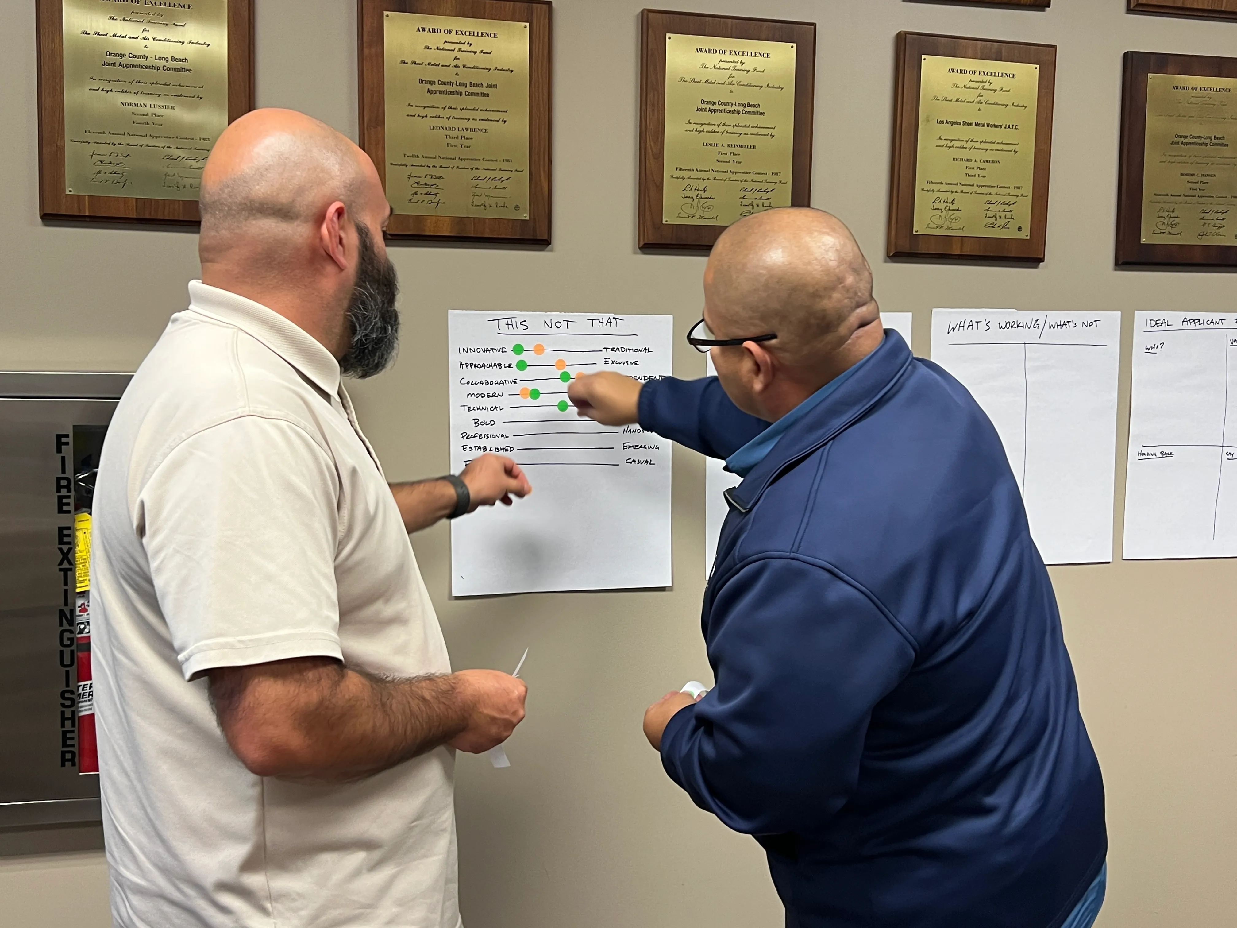 Two men discussing and pointing at charts on a wall with awards displayed above.
