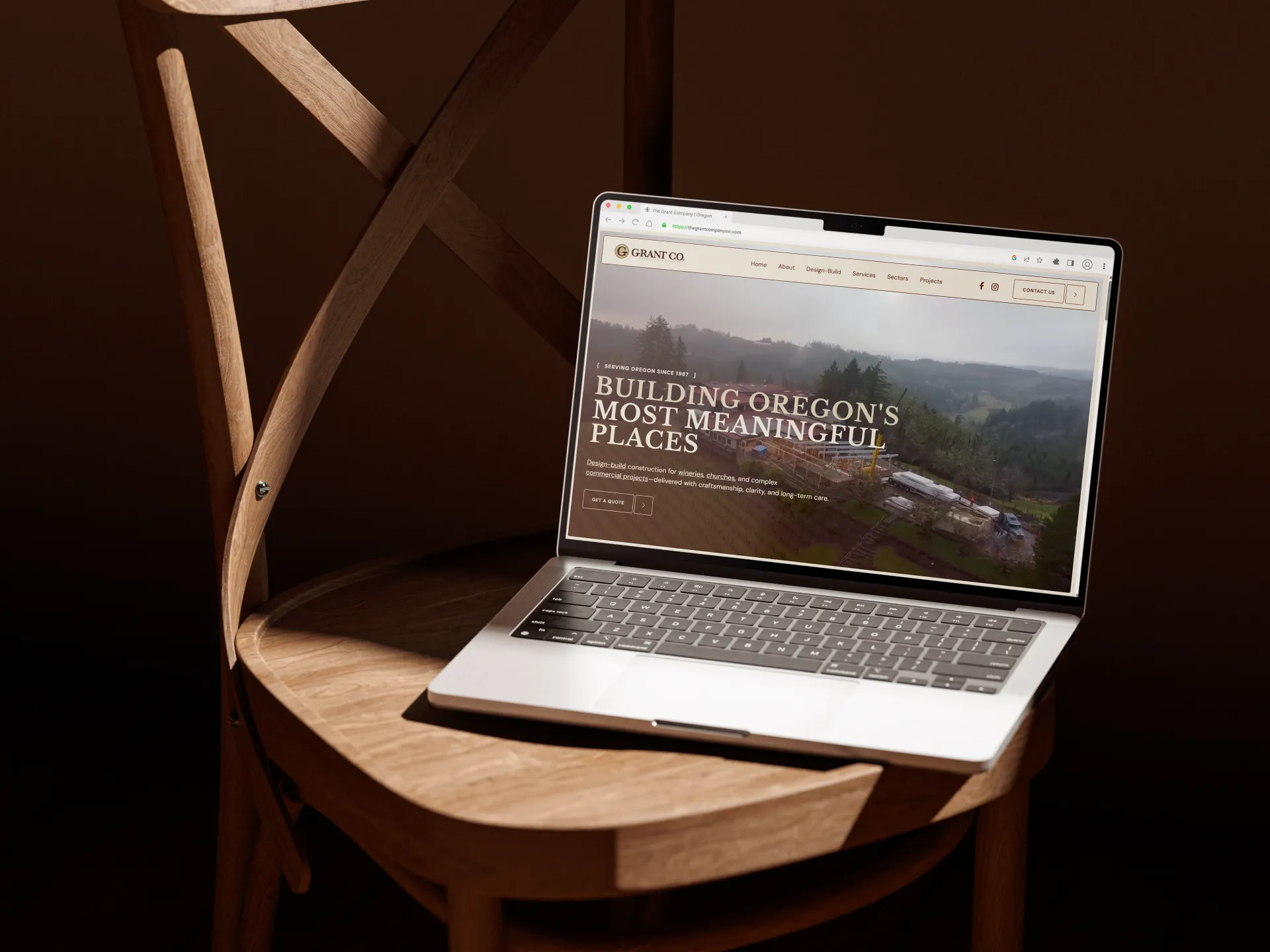 Laptop on wooden chair displaying The Grant Company website with text about building Oregon's meaningful places.