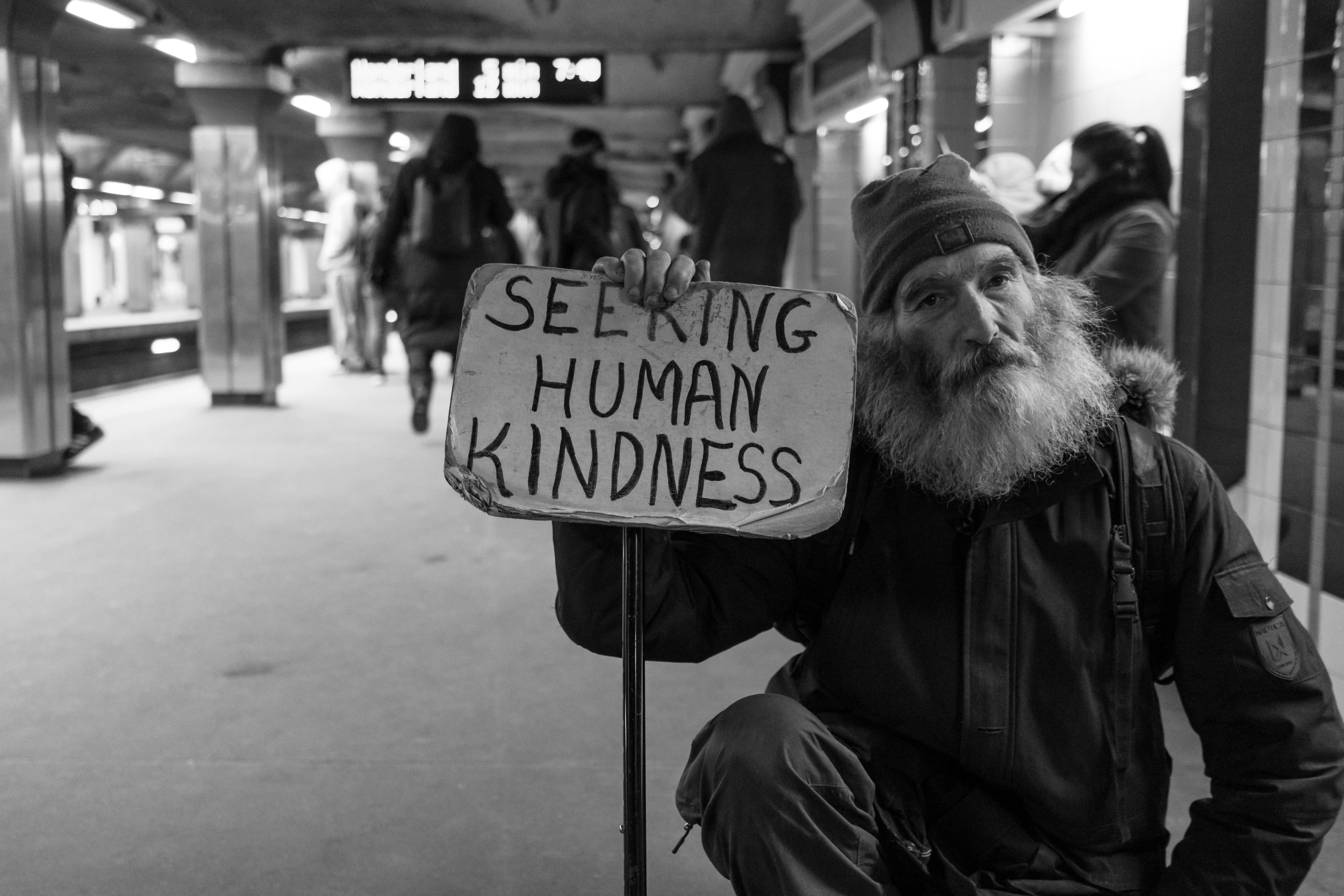 A bearded man in a subway station holding a sign that reads "Seeking human kindness"