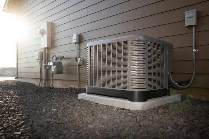 A residential air conditioning unit rests on a gravel bed next to a house, with utility meters and pipes visible.