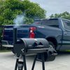 A black BBQ smoker emits smoke, set in a driveway beside a pickup truck under cloudy skies.