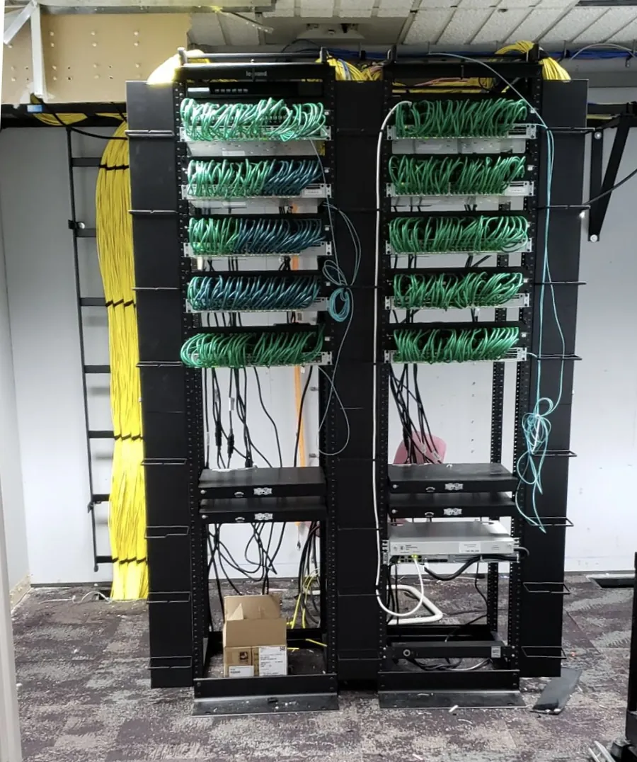 The same wiring closet after rework: two open racks with green patch cables neatly routed and dressed between the switches and patch panels.