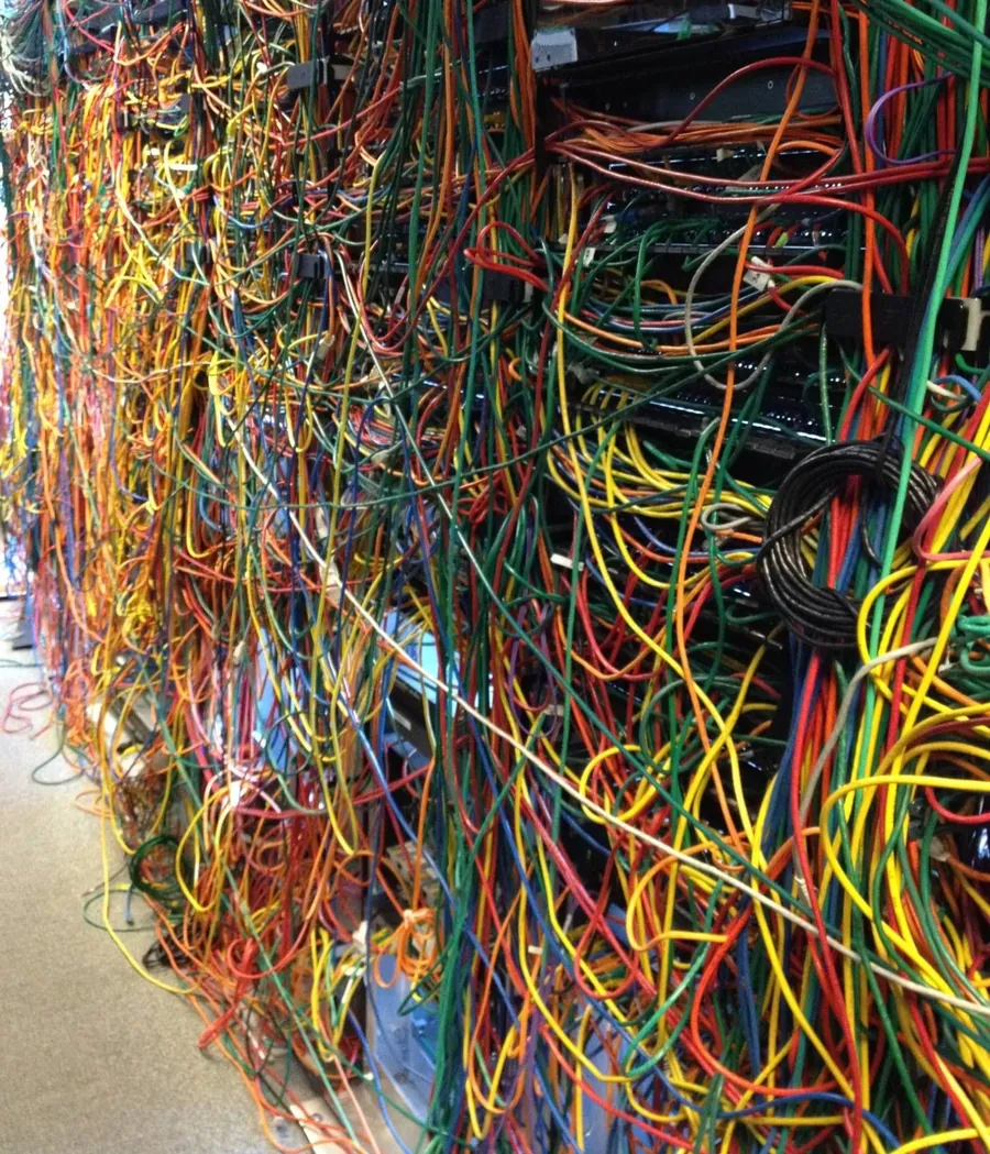 A business wiring closet before rework: hundreds of tangled red, yellow, green and blue network cables hanging loose across the switch racks, none of them labelled.