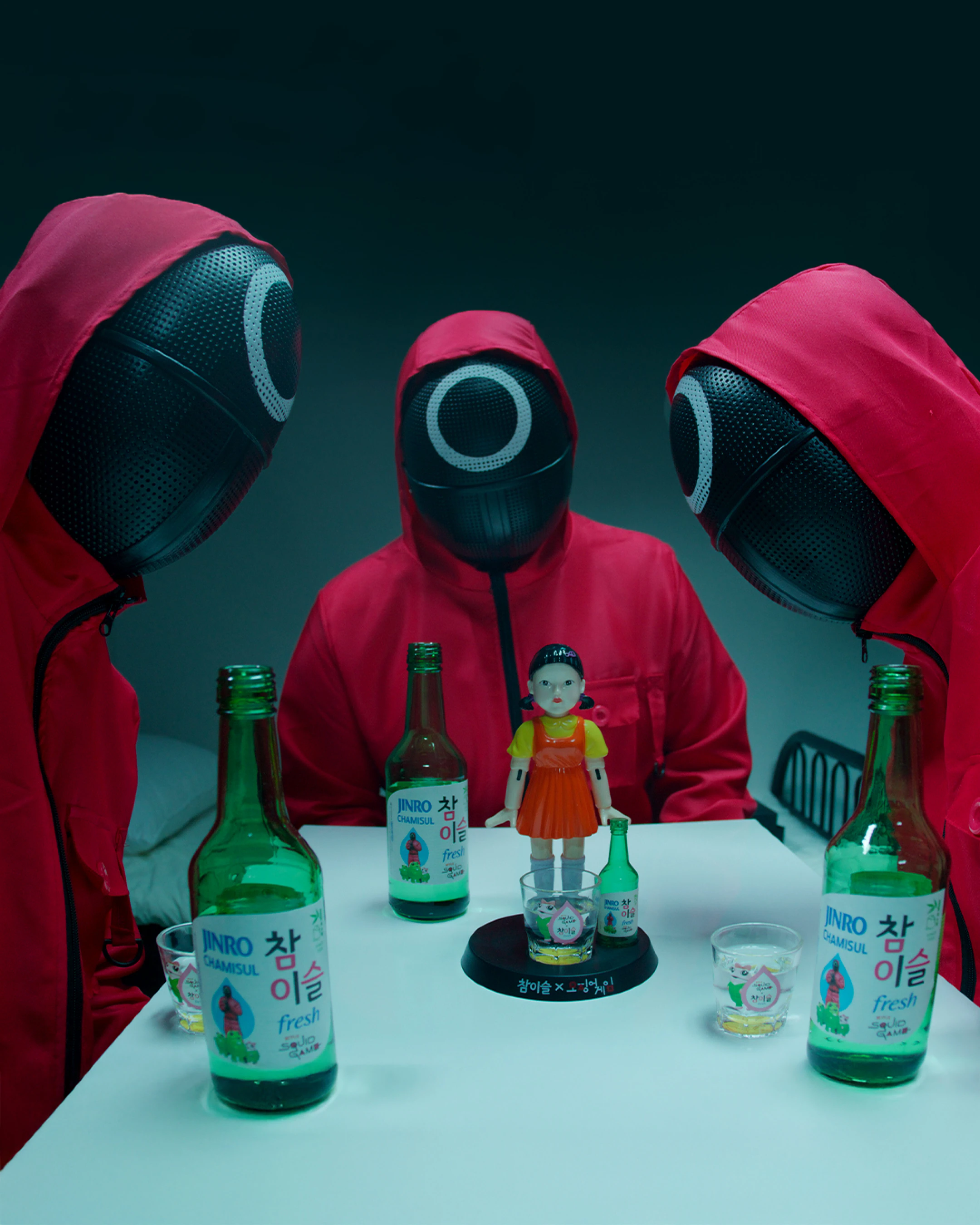 People in red hooded suits with masks sitting at a table with soju bottles and a doll figure
