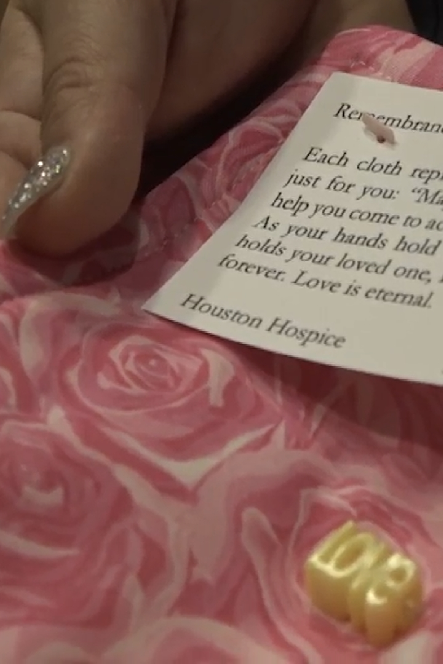 Close-up of a hand with glittery nail polish holding a pink fabric with a rose pattern, accompanied by a tag from Houston Hospice with a remembrance message and a small golden bead spelling 'Love.'