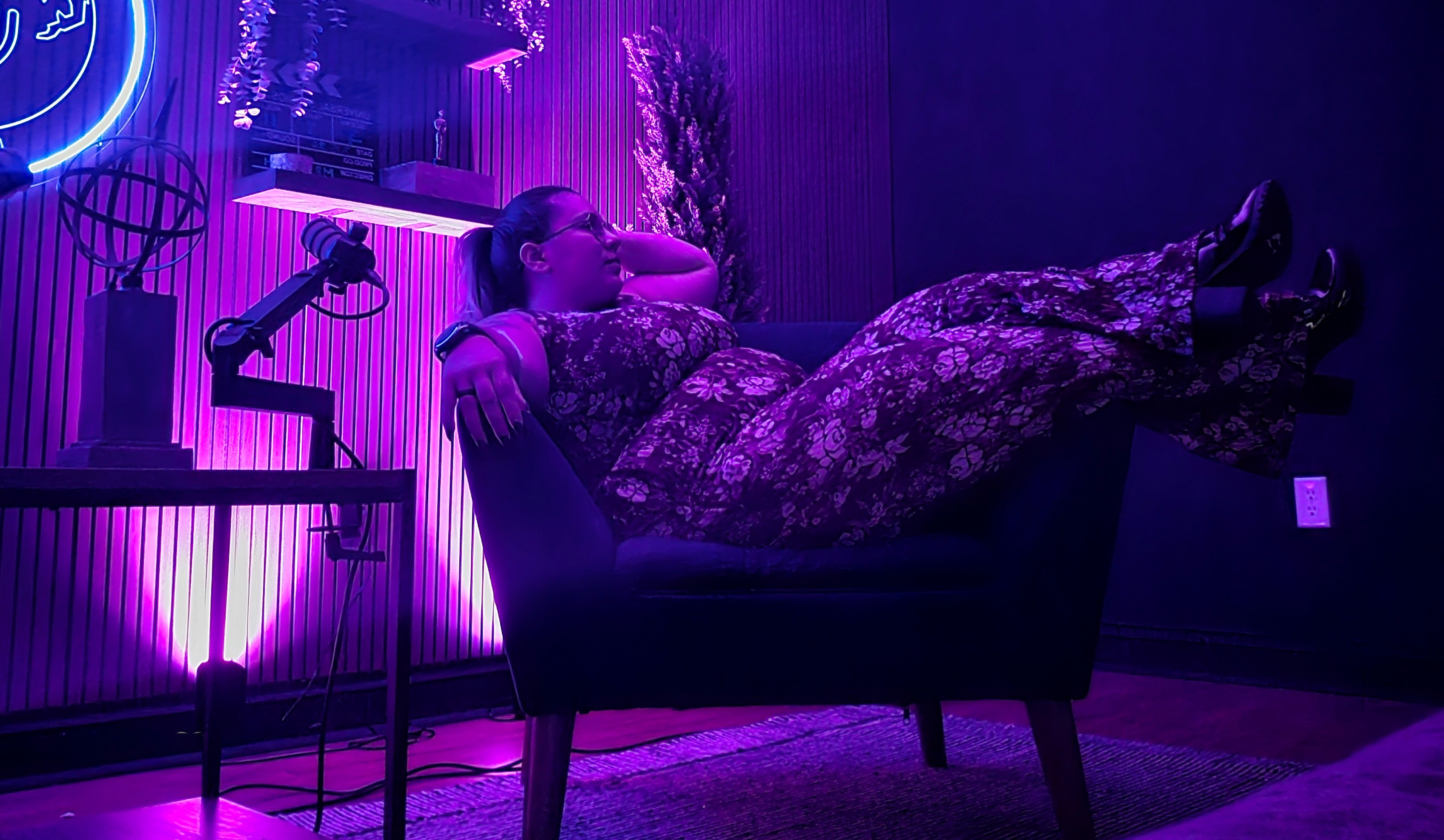 Woman wearing glasses and floral outfit reclining sideways on a dark chair with her legs resting on the armrest in a purple-lit room with a microphone and decorative elements in the background.