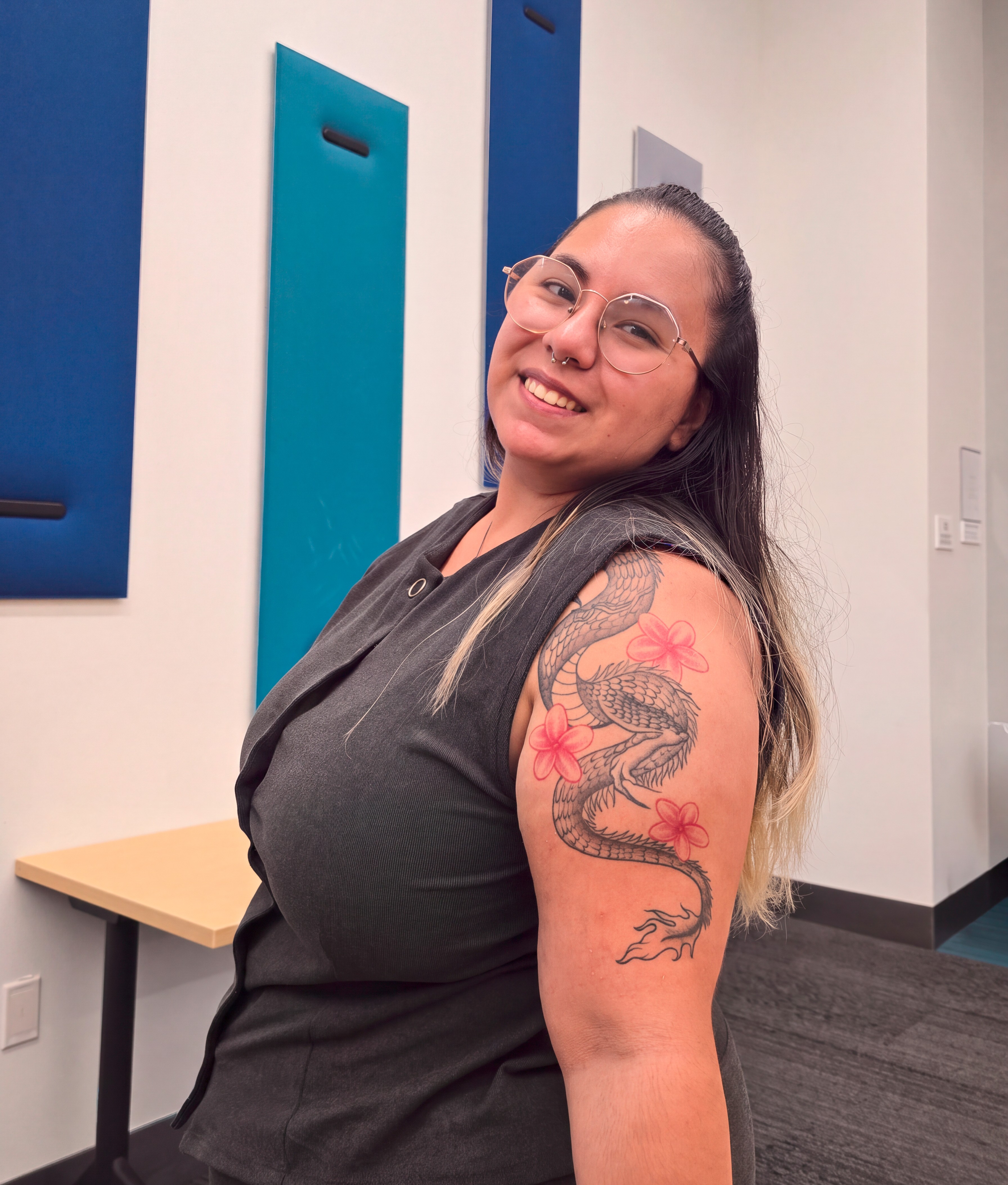Smiling woman with long dark hair and glasses showing a black and gray dragon tattoo with pink flowers on her upper arm, standing in a modern office space with blue wall panels and a wooden table.