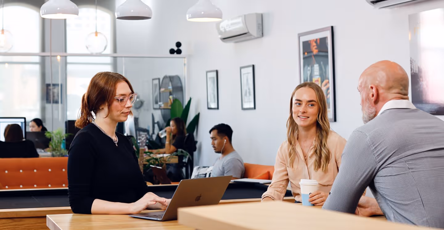 Three people engaging in a casual meeting at a modern office with laptops and coffee cups, with other employees working in the background.