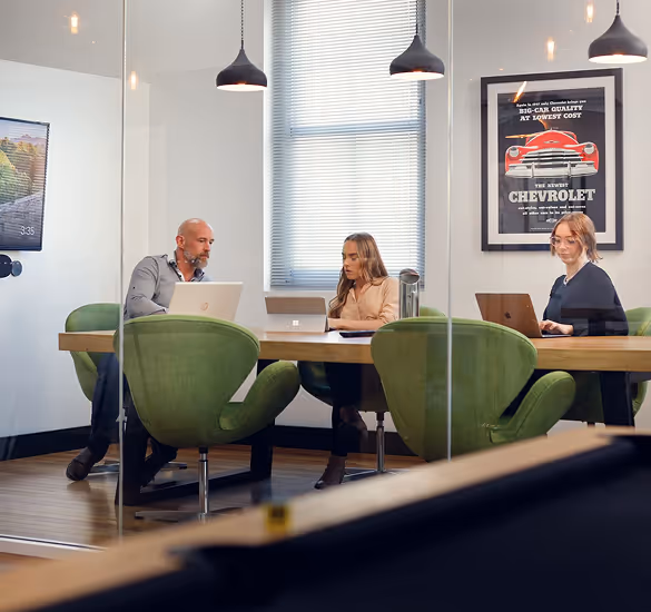 Three people working on laptops around a wooden table in a modern office with green chairs and a Chevrolet poster on the wall.