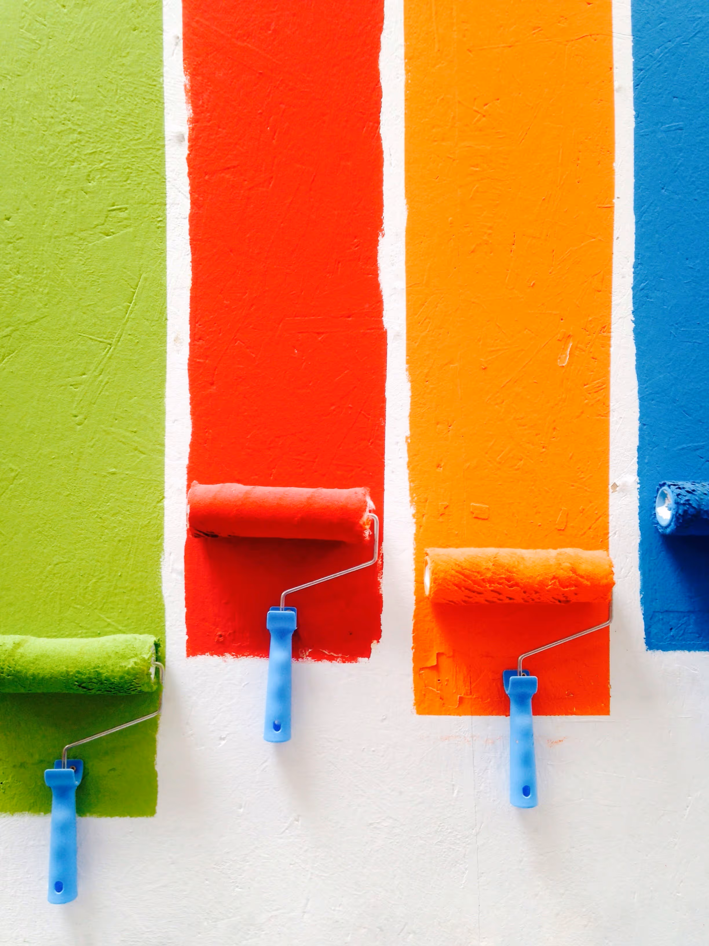 Four paint rollers with blue handles applying vertical stripes of green, red, orange, and blue paint on a white wall.