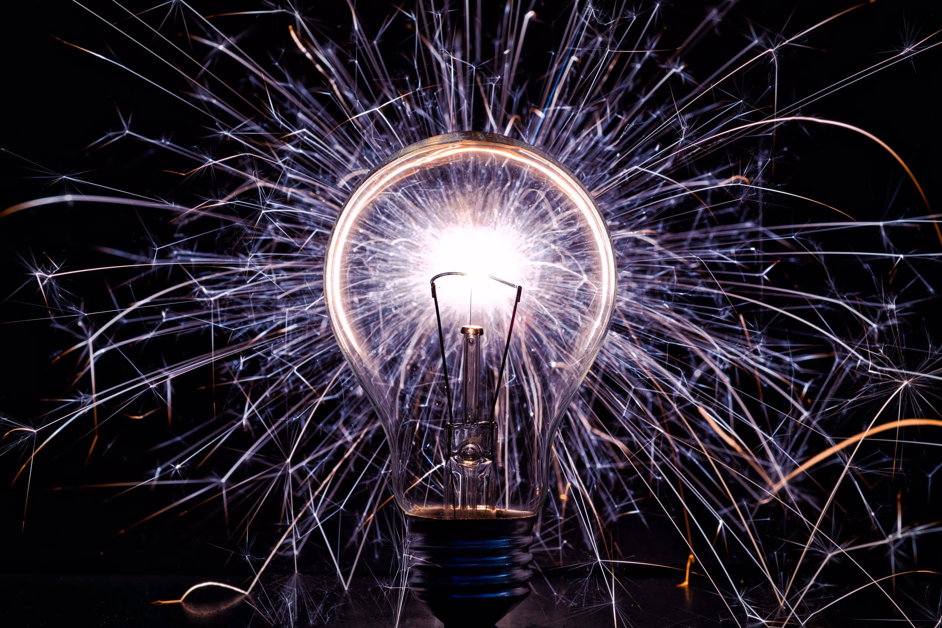 Close-up of a glowing incandescent light bulb with bright sparks radiating behind it on a black background.
