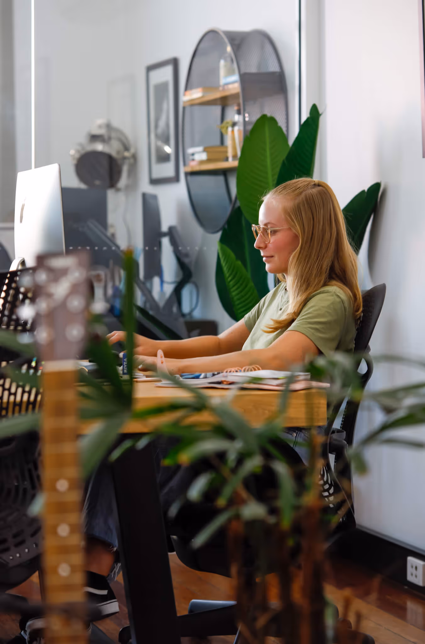 Young woman with glasses working on a computer at a wooden desk in a modern office with plants and shelves in the background.