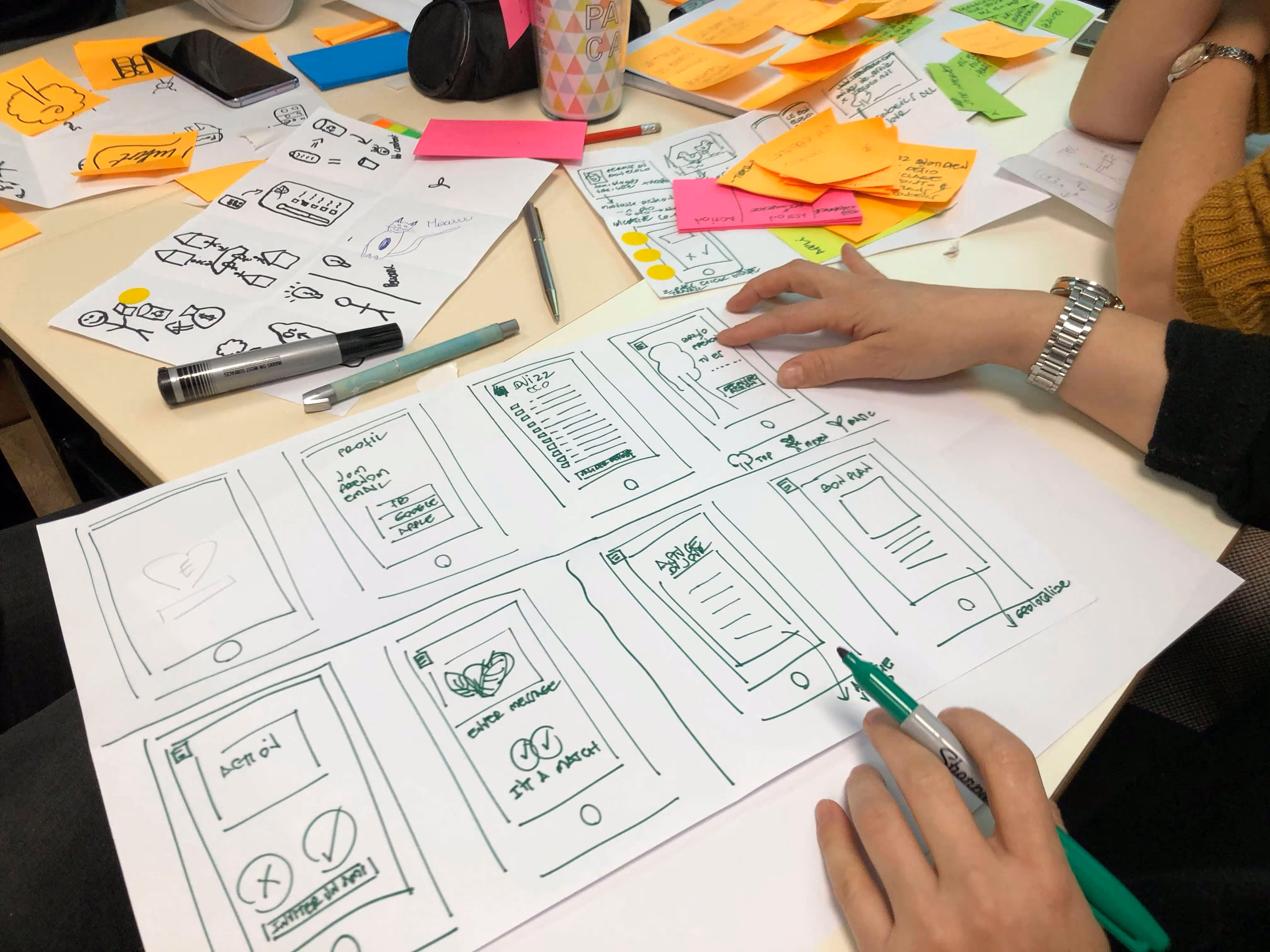 Person sketching mobile app wireframes on large sheets of paper with various sticky notes and pens scattered on the table.