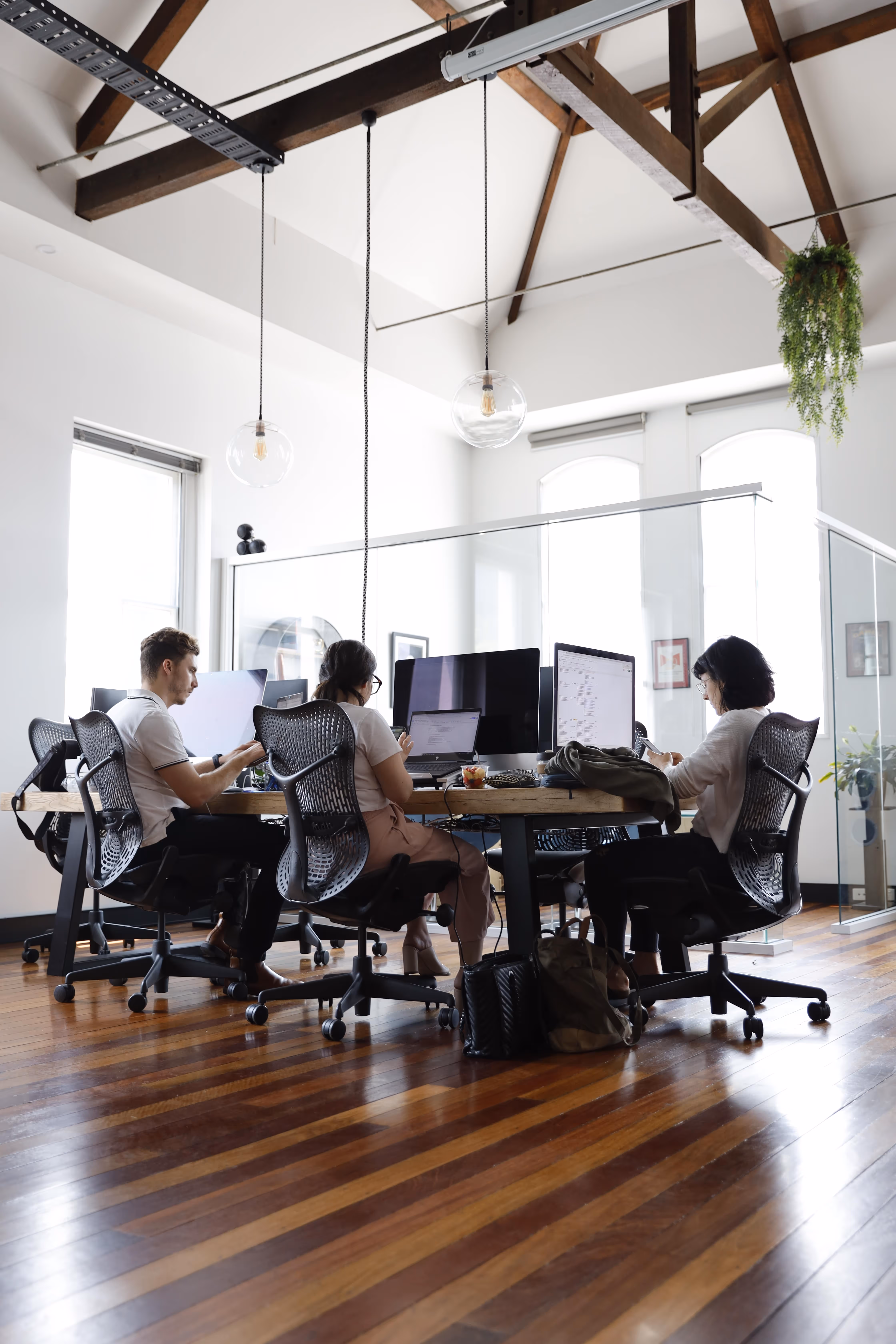 Three people seated at a wooden table in a bright office working on computers under exposed wooden ceiling beams.