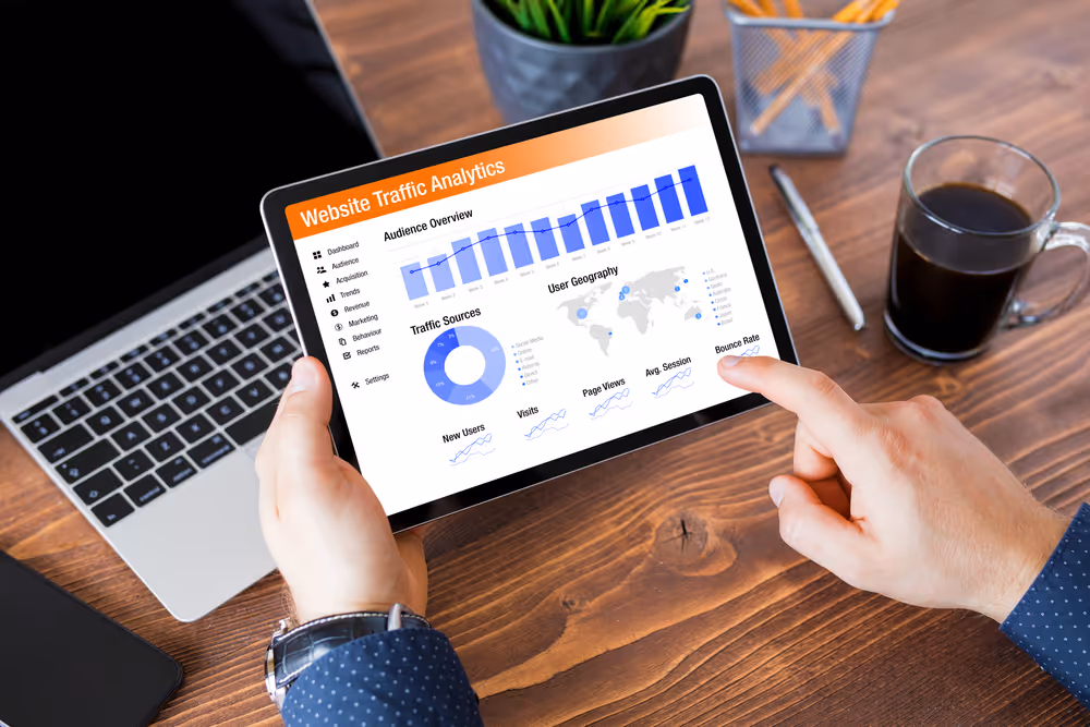 Person holding a tablet displaying website traffic analytics with graphs and charts on a wooden desk near a laptop, coffee, and pencil holder.