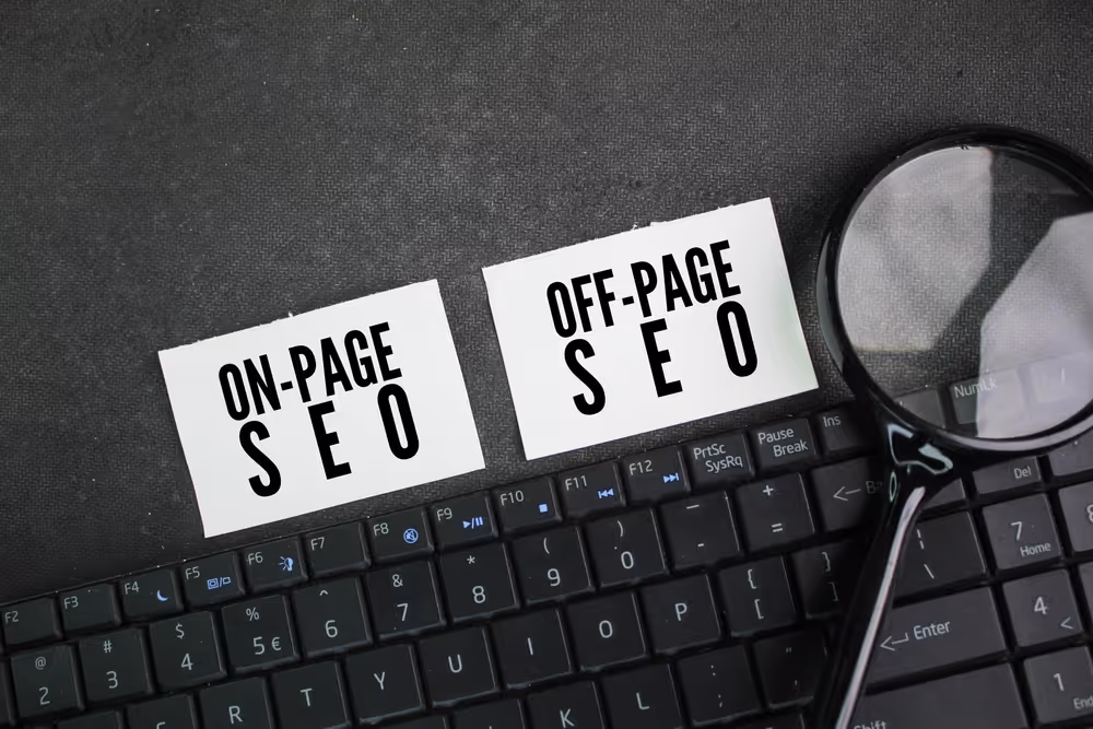Two white cards labeled 'ON-PAGE SEO' and 'OFF-PAGE SEO' placed on a black keyboard with a magnifying glass nearby.
