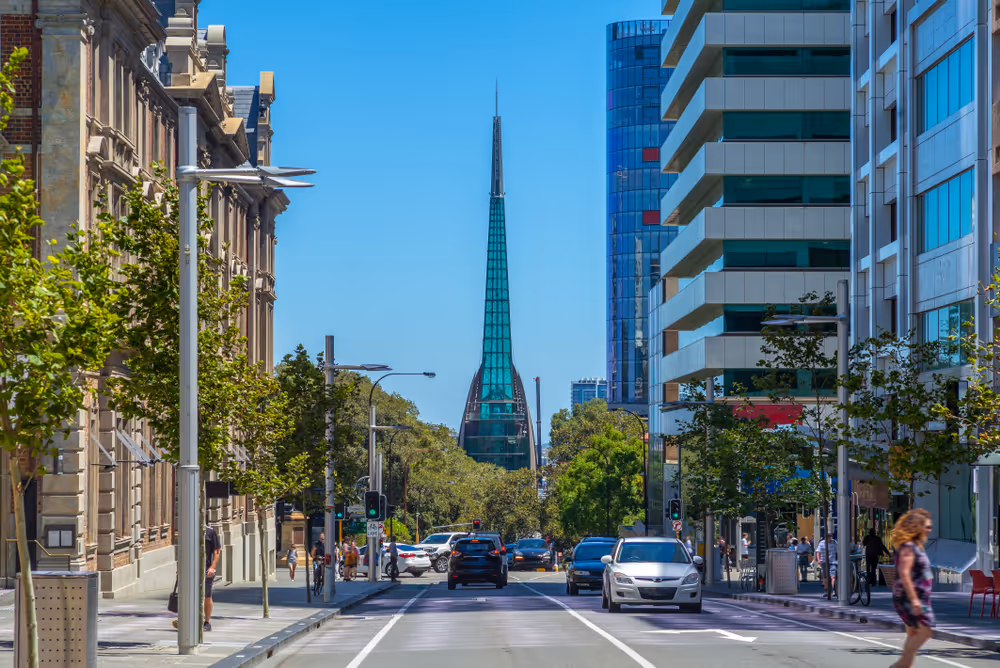 Urban street view with cars, pedestrians, trees, and the glass spire of a modern building in the background under a clear blue sky.