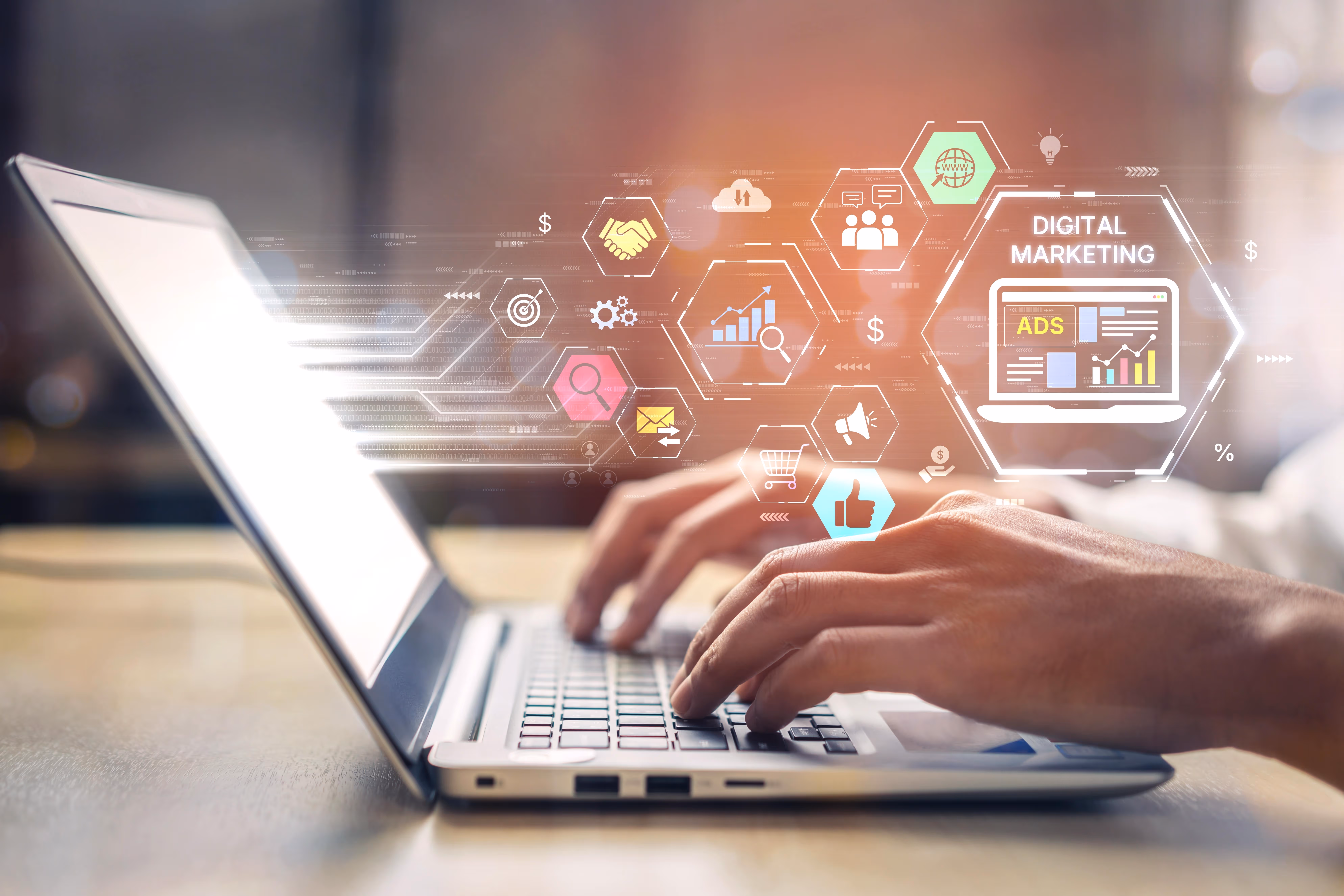 Hands typing on a laptop keyboard with digital marketing and advertising icons floating above, including charts, a shopping cart, a globe, and a thumbs-up symbol.