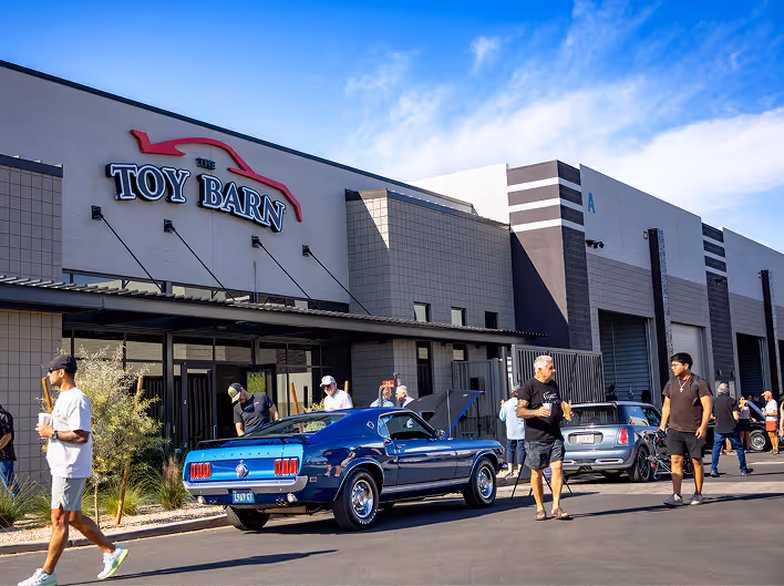 Classic blue Ford Mustang parked outside a building with people walking nearby under a blue sky.