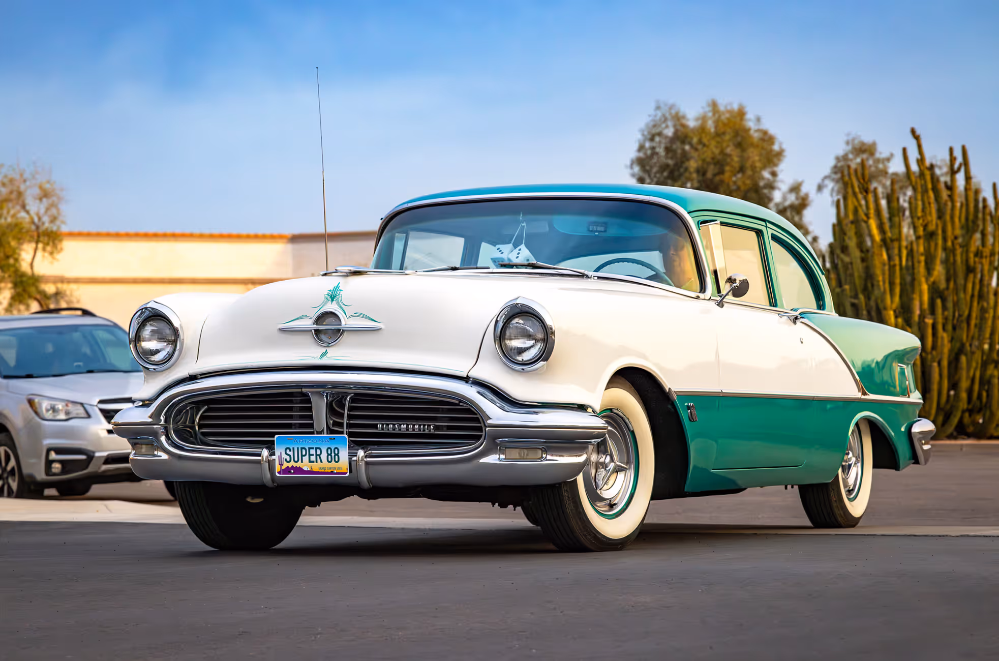 Green and white vintage Oldsmobile car parked on street with a cactus and other cars in the background.