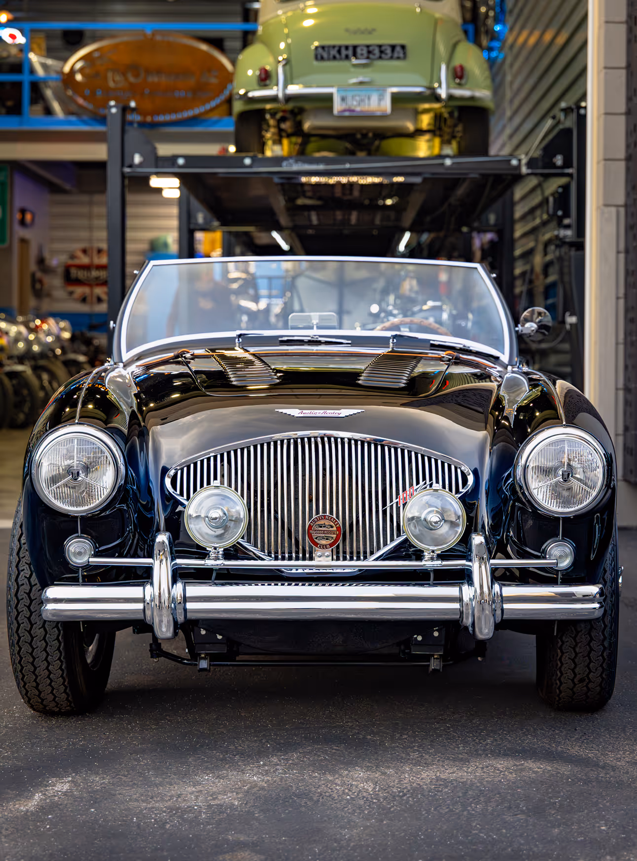 Front view of a shiny black vintage convertible car inside a garage with another green vintage car elevated behind it.
