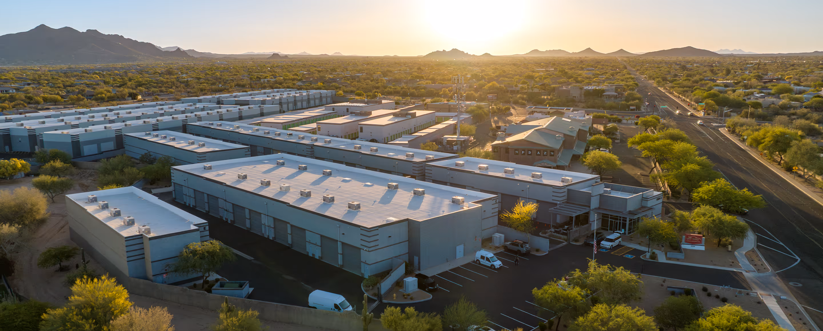 Aerial view of a modern industrial complex with multiple white-roofed buildings and parking lots surrounded by green trees and distant mountains at sunset.