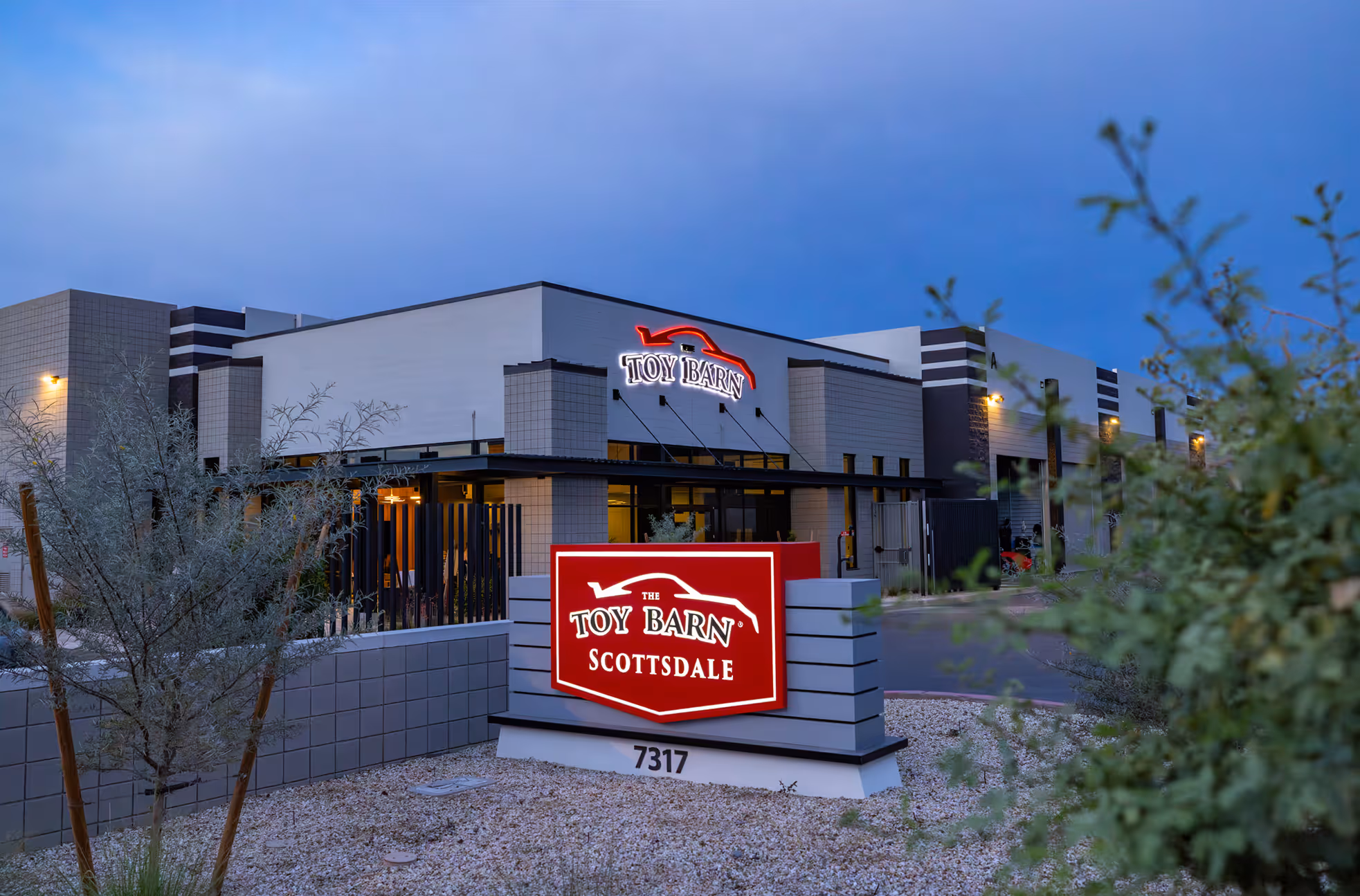 Exterior view of Toy Barn Scottsdale sign illuminated at dusk with greenery around.
