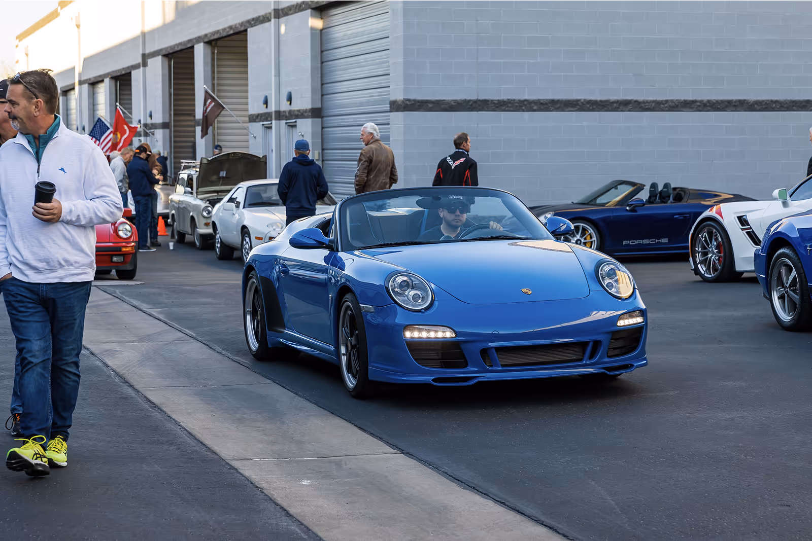 Blue Porsche convertible sports car driving past parked cars and people outside a garage building.