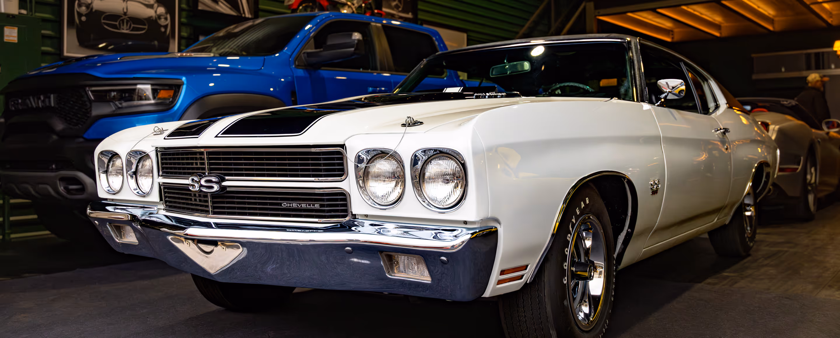 White Chevrolet Chevelle SS classic muscle car parked indoors next to a blue Ram truck.