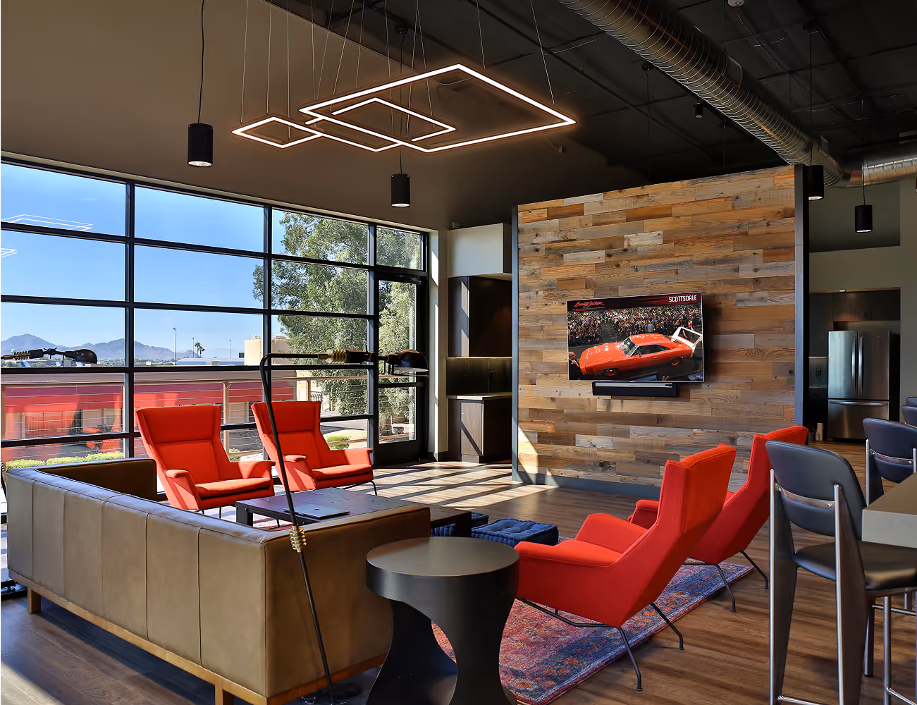 Modern lounge area with large windows, red armchairs, beige sofa, mounted TV, and wooden accent wall.