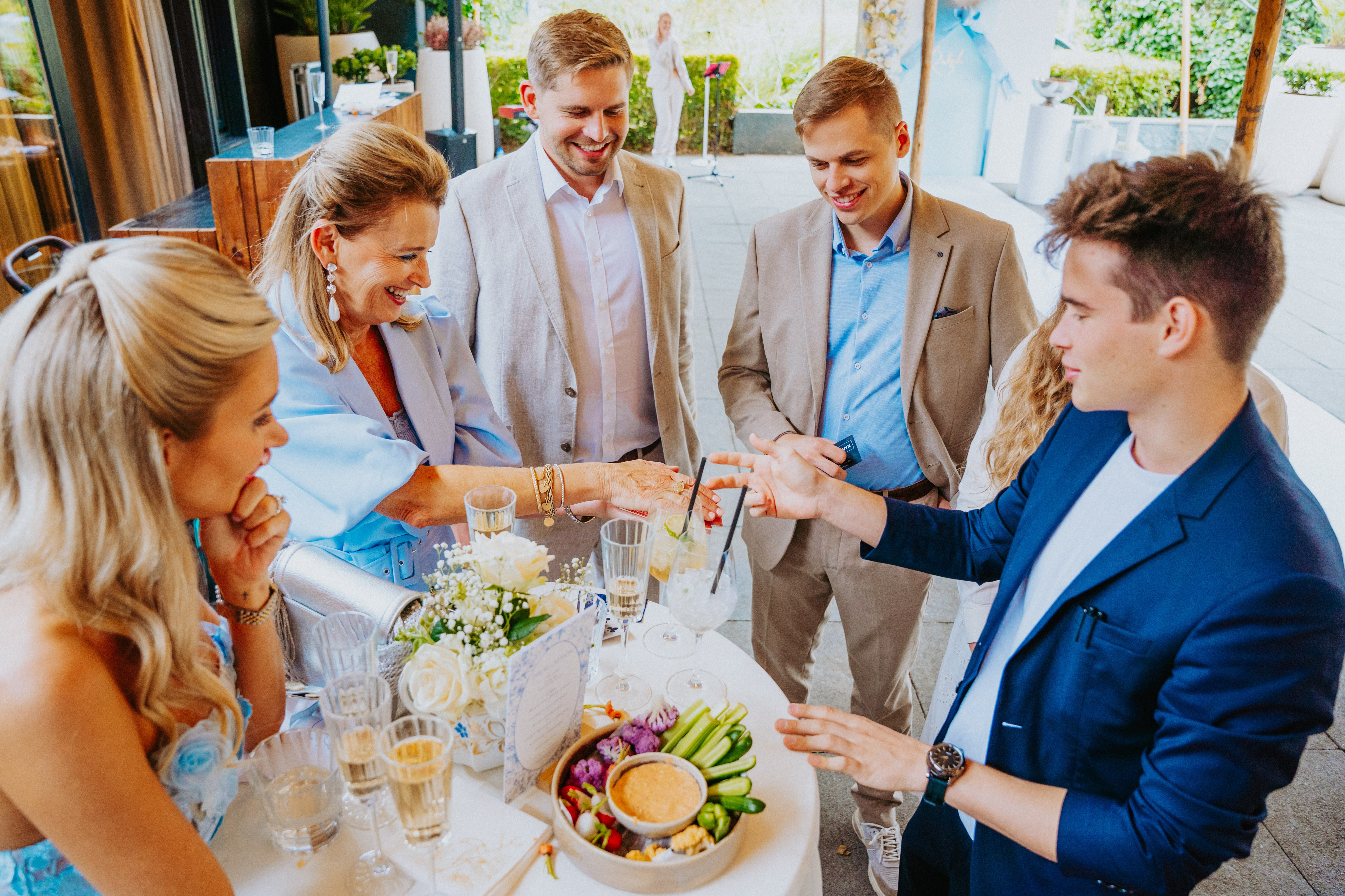 Interactie tussen goochelaar Jonas De Bruyn en gasten, magische truc tijdens chic feest met hapjes en drankjes, focus op entertainment en sfeervolle setting