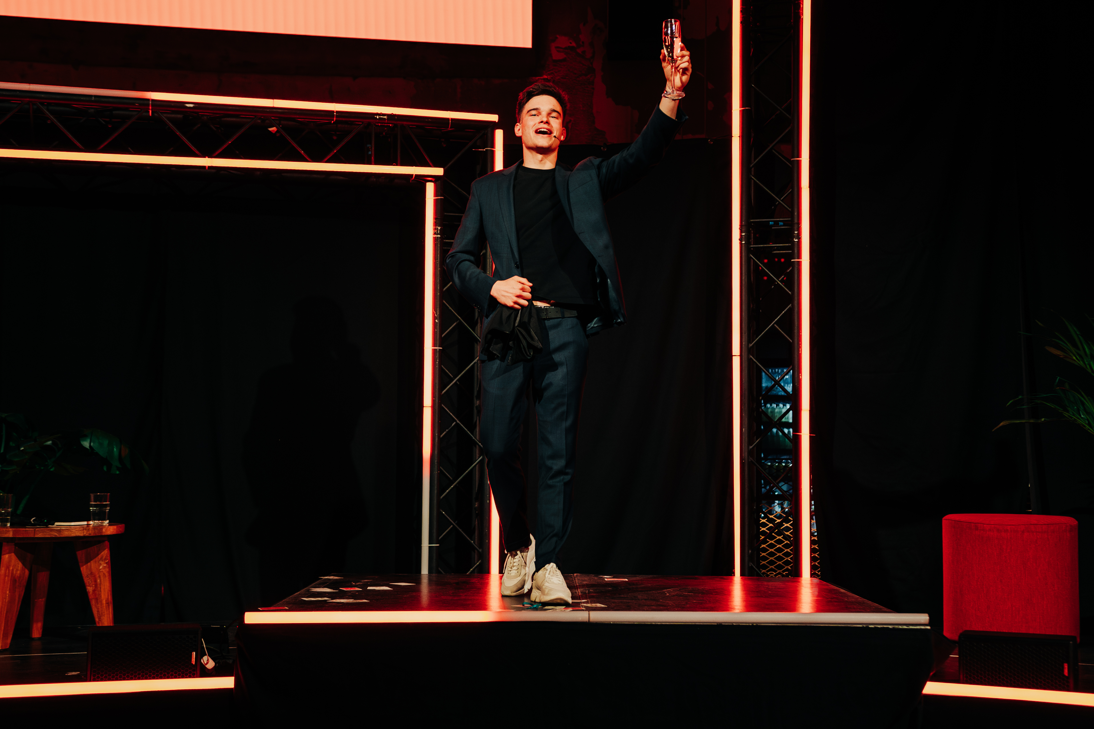 Man in donkerblauw kostuum op podium tijdens business event, houdt glas omhoog als toost, moderne verlichting en industriële setting