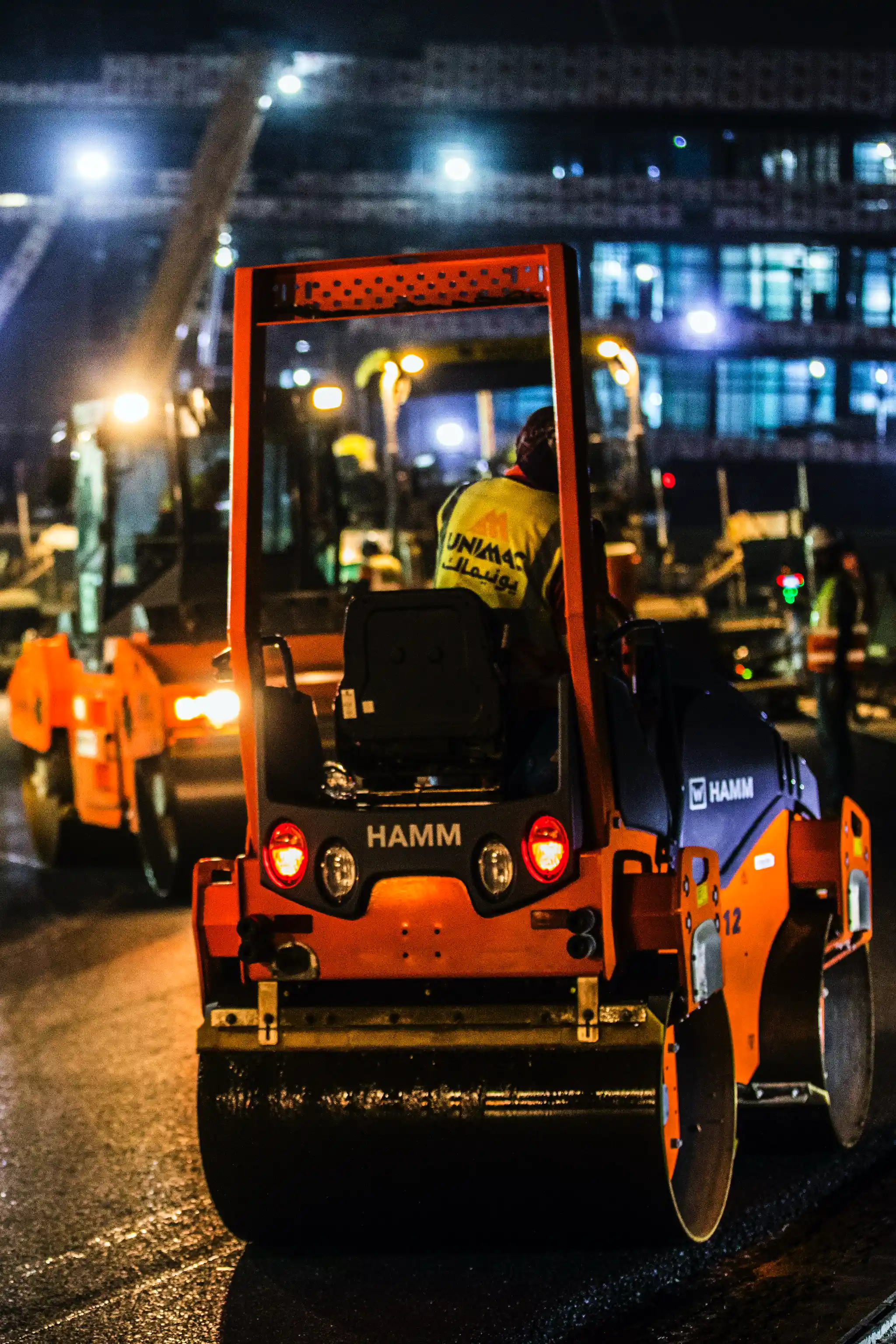 Construction worker operating a HAMM road roller machine at night on a construction site.