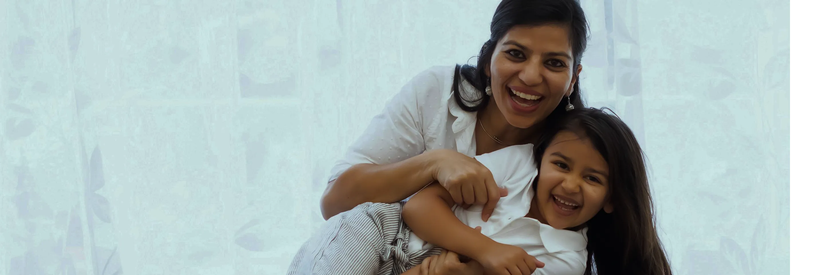 Smiling woman hugging a laughing young girl with long hair in front of a light curtain.