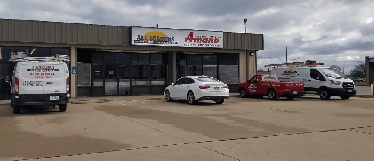 Parking lot with three All Seasons company vehicles—two vans and one red pickup truck—in front of a commercial building under cloudy sky.