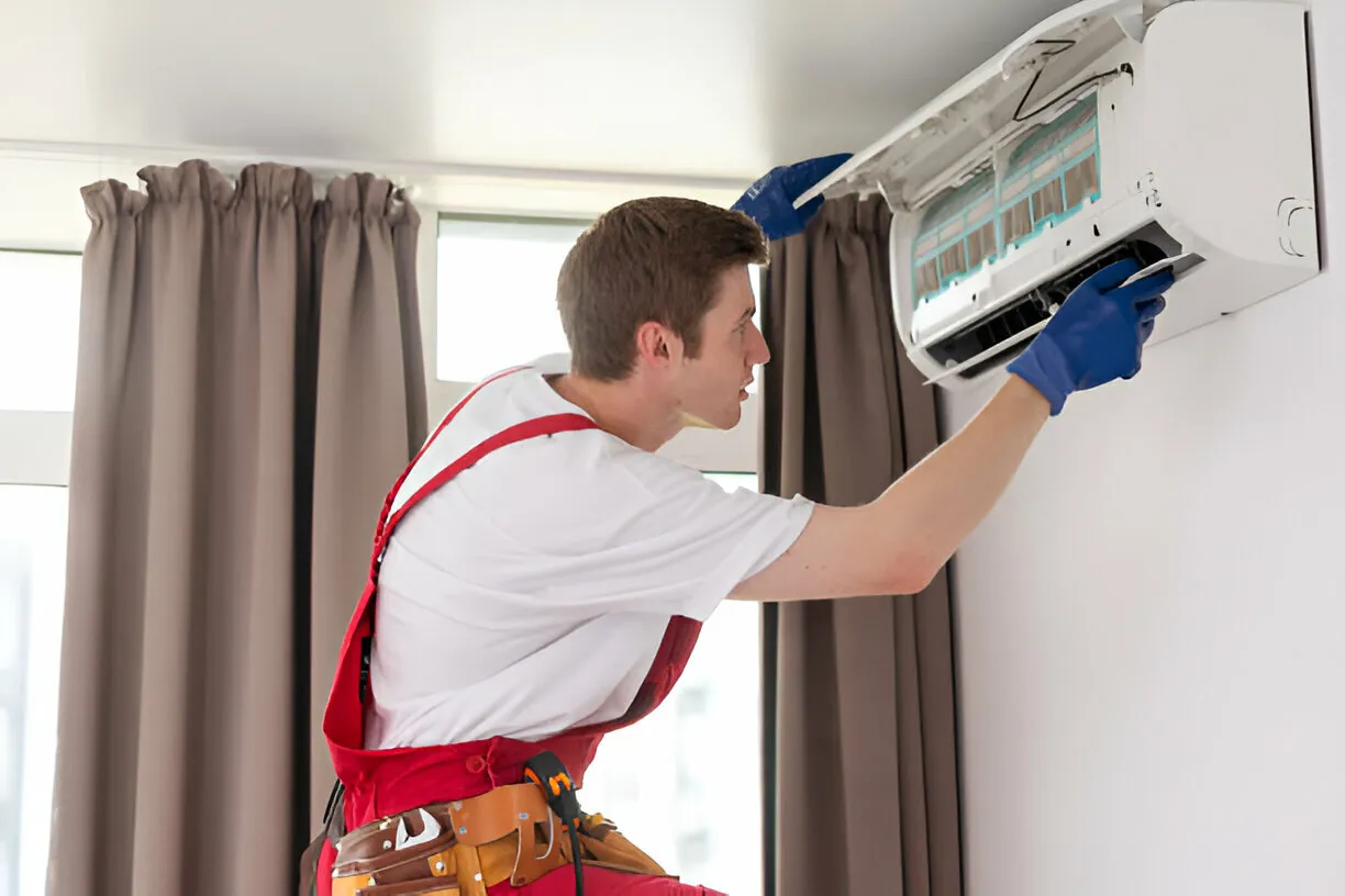 Technician wearing gloves and red overalls servicing a wall-mounted air conditioner inside a room.