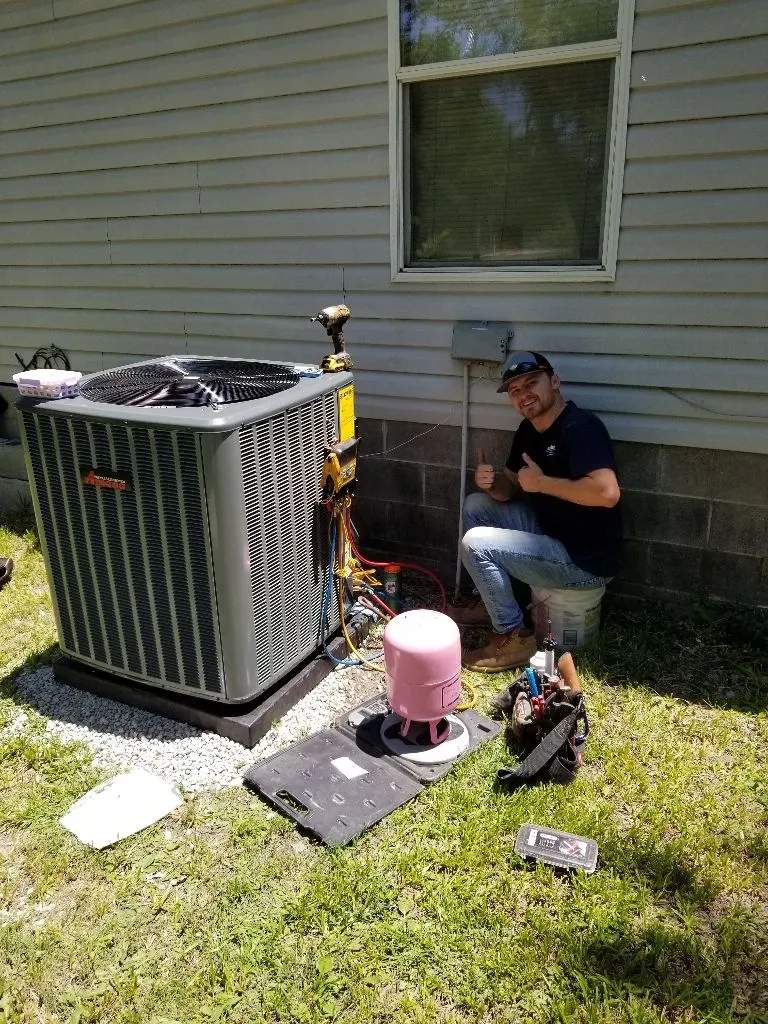 Technician sitting on a bucket thumbs up next to an HVAC unit outside a house with repair tools and equipment.