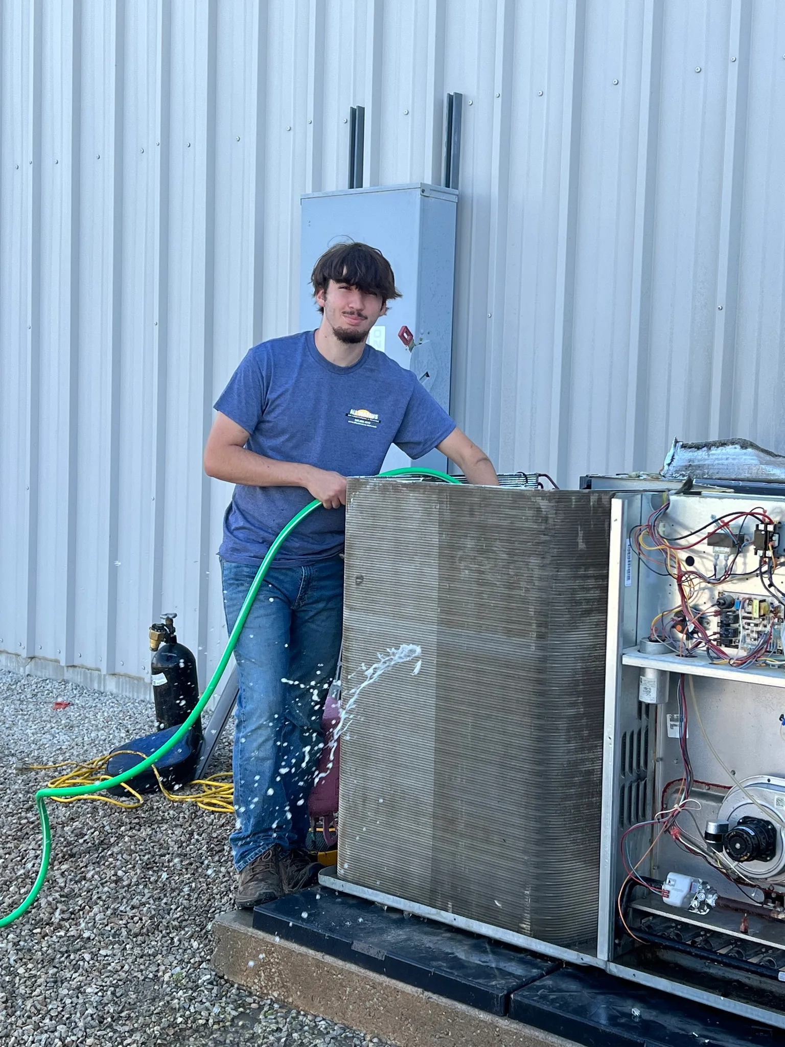 Technician holding a green hose spraying water on an outdoor HVAC unit's condenser coils beside a metal building wall.