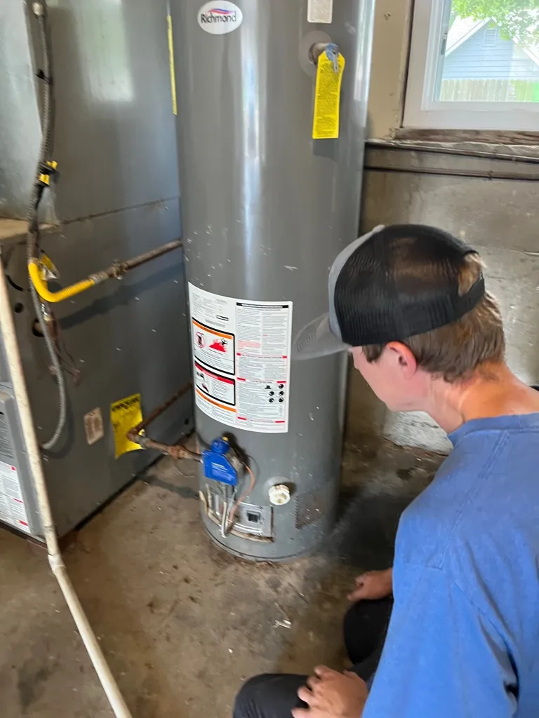 Young man in a blue shirt and black cap inspecting a gray Richmond water heater in a basement.