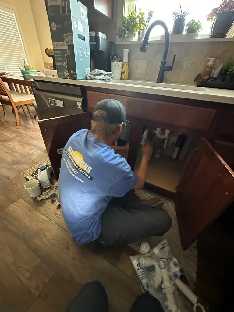 A plumber in a blue shirt and cap kneeling and repairing pipes under a kitchen sink with tools and plumbing parts nearby.
