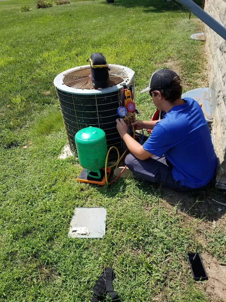 Technician in a blue shirt and cap working on an air conditioning unit outdoors with gauges.