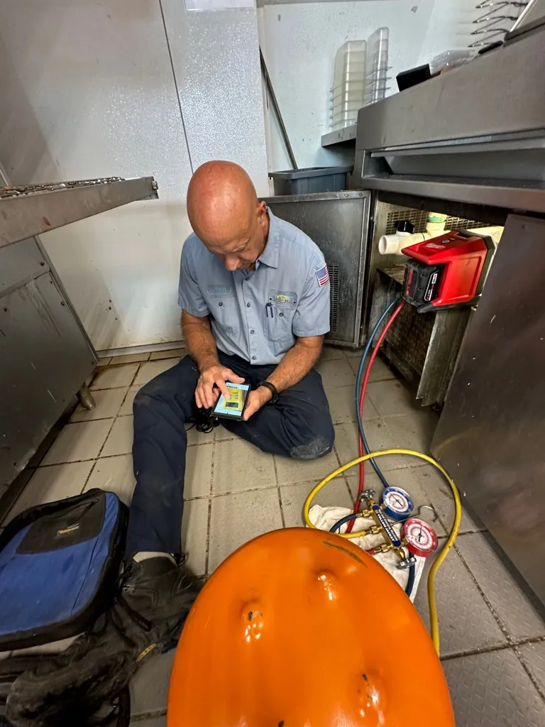 Technician sitting on a tiled floor using a handheld device while working on commercial refrigeration equipment with gauges and hoses connected.