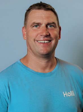 Smiling man with short brown hair wearing a light blue t-shirt with the word 'Holli' on it against a plain light blue background.