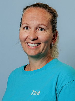 Smiling woman with light skin and hair tied back wearing a turquoise shirt with the name 'Tina' on it, against a light blue background.