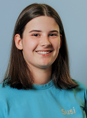 Smiling young woman with straight brown hair wearing a turquoise shirt with the name 'Susi' embroidered on it, against a plain light blue background.