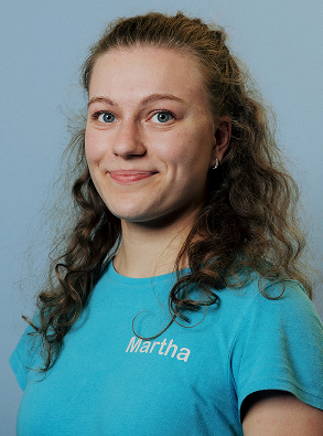 Young woman with curly hair wearing a blue shirt with the name Martha on it smiling against a blue background.